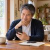 自宅でお茶を飲みながらスマートフォンを見つめ、プロフィールを考える50代男性のイメージ